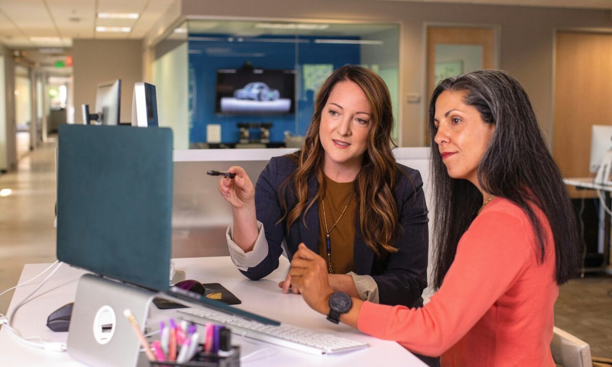 Two women stand at a desk in an office looking at a desktop computer, with one pointing at the screen