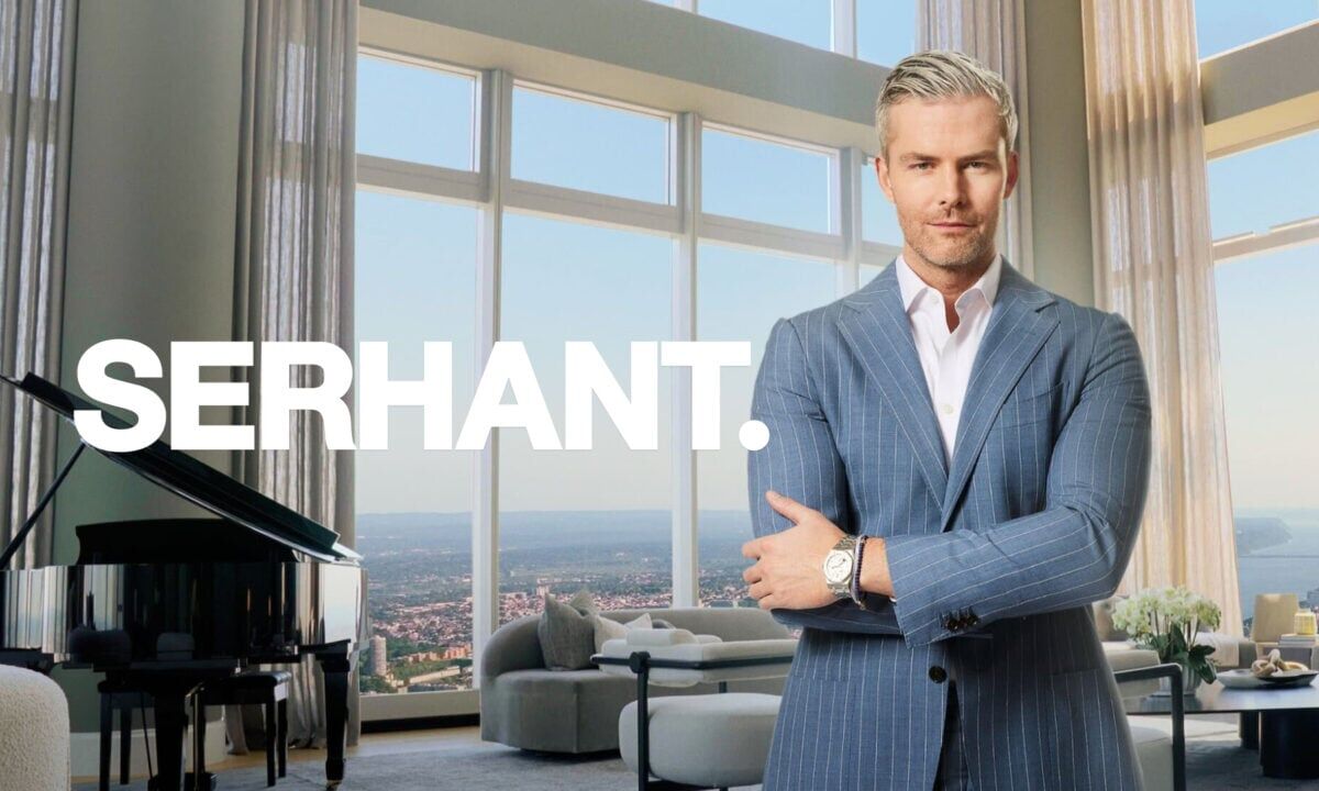 Ryan Serhant stands with crossed arms in a luxury high-rise with his brokerage's logo to his left