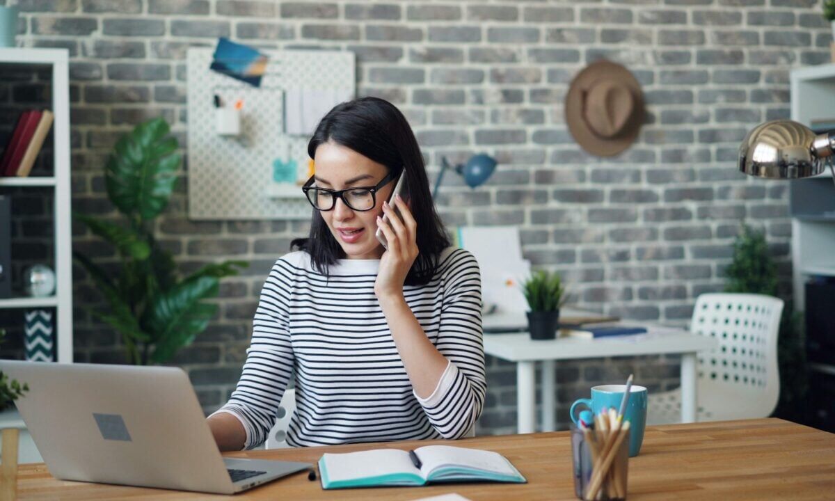 A real estate agent sits at a desk in front of a laptop on her phone, trying out real estate lead generation ideas to increase business
