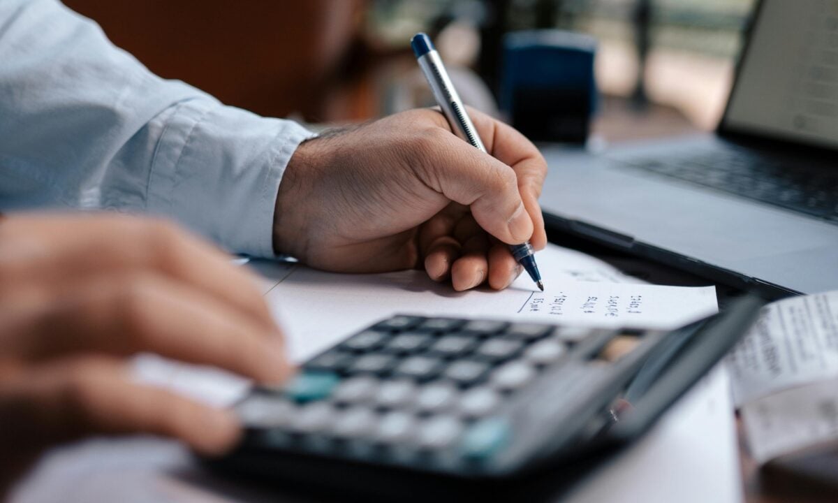 A real estate agent uses a calculator, laptop and pen and paper to figure out their email marketing costs